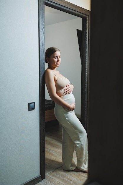 Cute young pregnant woman in a top and casual maternity clothes Cute pregnant woman hugging her belly at home