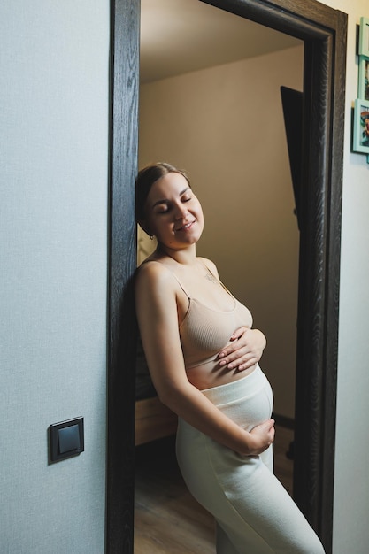 Cute young pregnant woman in a top and casual maternity clothes Cute pregnant woman hugging her belly at home