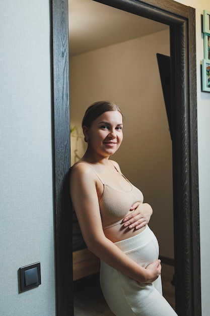 Cute young pregnant woman in a top and casual maternity clothes Cute pregnant woman hugging her belly at home