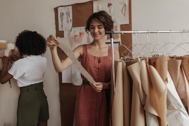 Cute young shorthaired woman in linen red dress looks at clothes pattern Curly brunette darkskinned lady designs dress