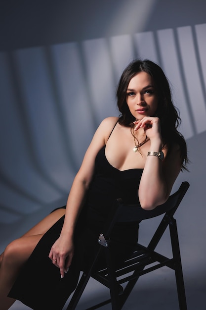 A cute young woman in a beautiful black dress sits on a chair and poses in the studio An attractive woman wears a black dress with a slit The girl looks sensual Summer casual street look