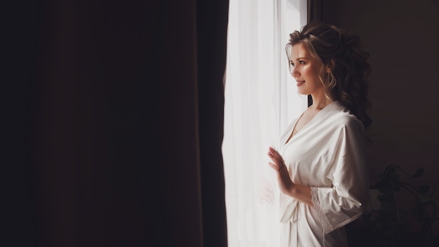 Cute young woman bride in home bathrobe at window in hotel room. Morning of bride on wedding day