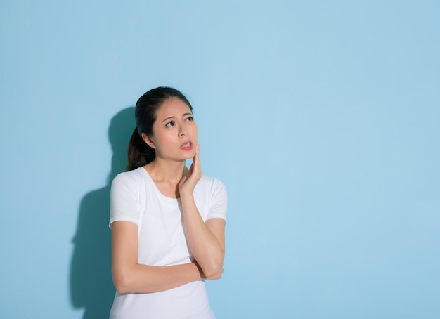 cute young woman holding on chin performance tooth painful uncomfortable emotion and looking at empty area standing in blue wall background.