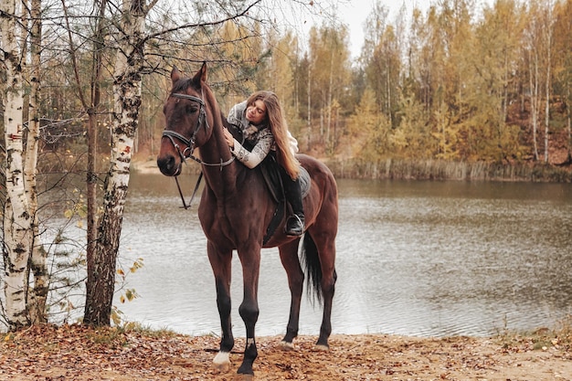 Cute young woman on horse in autumn forest by lake