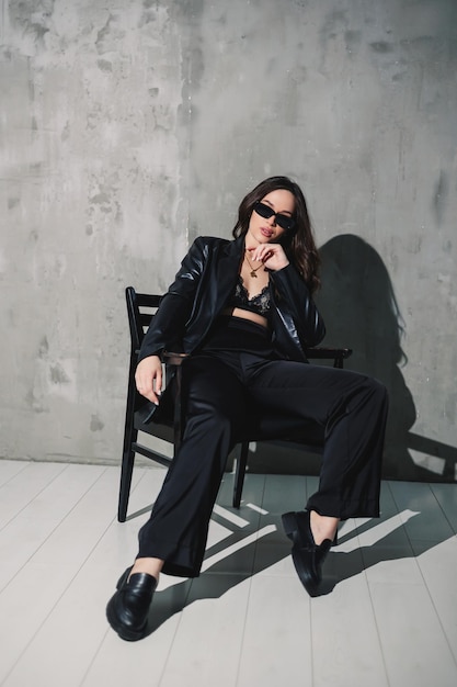 A cute young woman in a leather jacket and black pants wearing sunglasses is sitting on a chair on a gray background