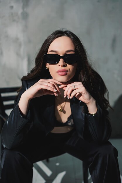 A cute young woman in a leather jacket and black pants wearing sunglasses is sitting on a chair on a gray background