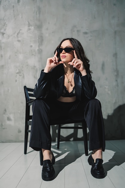 A cute young woman in a leather jacket and black pants wearing sunglasses is sitting on a chair on a gray background