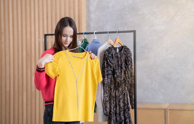 Cute young woman standing in front of hanger rack and trying to choose outfit dressing for work or walk Selection of a wardrobe stylist shopping