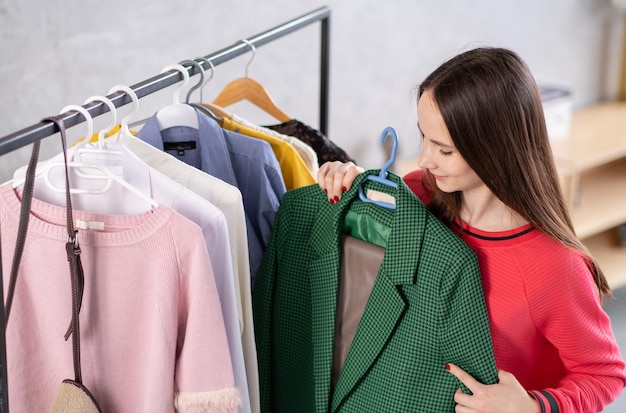 Cute young woman standing in front of hanger rack and trying to choose outfit dressing for work or walk Selection of a wardrobe stylist shopping