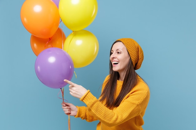 Cute young woman in sweater hat posing isolated on blue background. Birthday holiday party people emotions concept. Mock up copy space. Celebrating hold colorful air balloons point index finger aside.