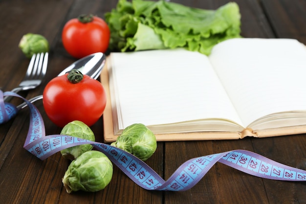 Cutlery tied with measuring tape and book with vegetables on wooden background