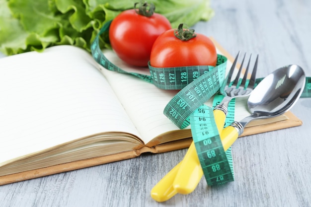 Cutlery tied with measuring tape and book with vegetables on wooden background