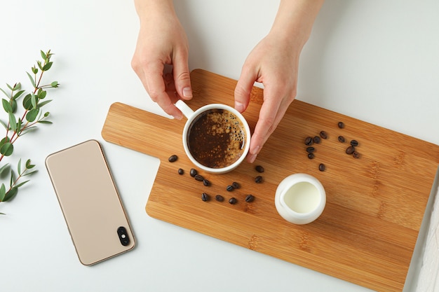 Cutting board with cup of coffee, coffee beans, phone and plant branch on white background