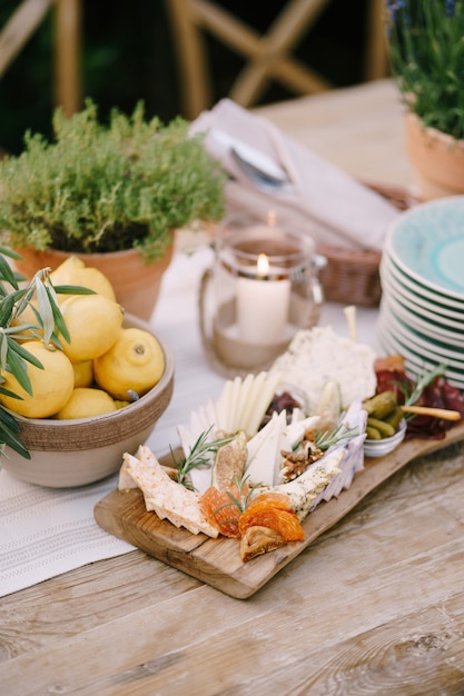 A cutting board with a variety of cheeses on a table with clean plates lemon and potted flowers