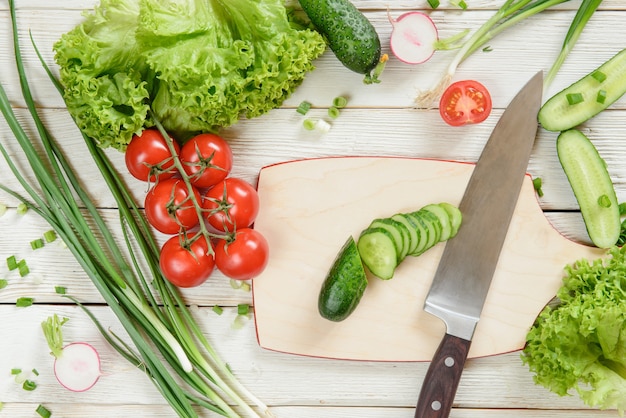 Cutting cucumber and fresh ingredients (tomatoes, lettuce, onion) for the vegetable salad on white wooden background, top view