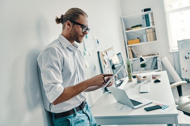 Cutting edge technologies. Good looking young man in shirt using digital tablet while standing in the office