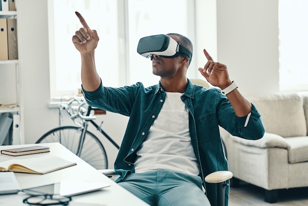 Cutting edge technologies. Handsome young African man wearing virtual reality simulator headset while sitting indoors