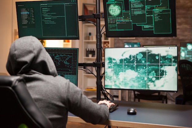 Cyber criminal wearing a hoodie writing a dangerous virus using computer with multiple screens.