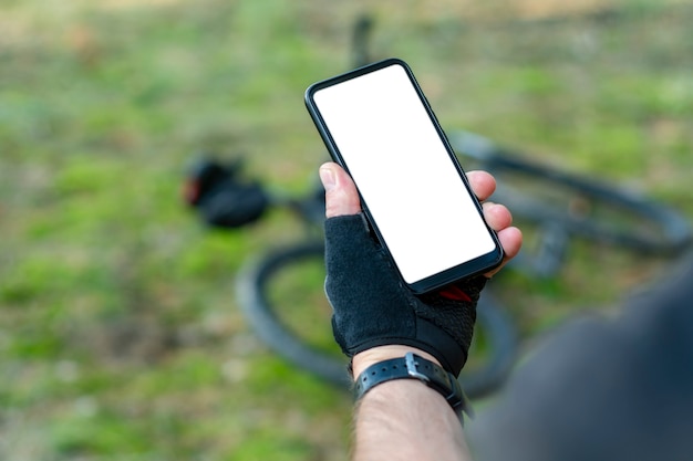 Cyclist holds mockup smartphone close-up with a white screen in his hands on the background of a bicycle in nature.