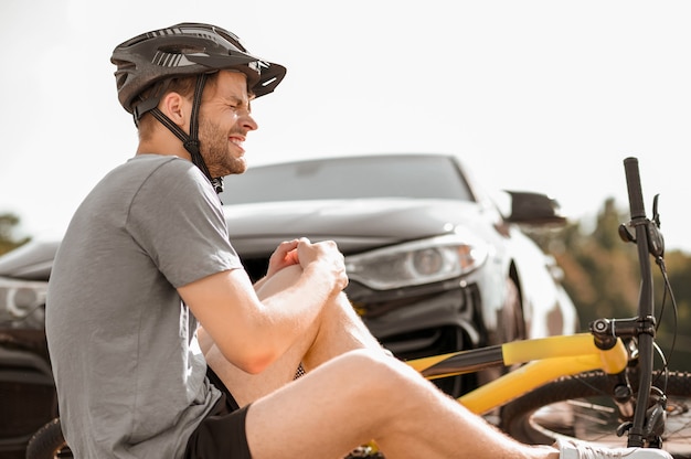 Cyclist, injury. Profile of unhappy young adult cyclist with knee injury while falling while sitting on ground in front of car on summer day