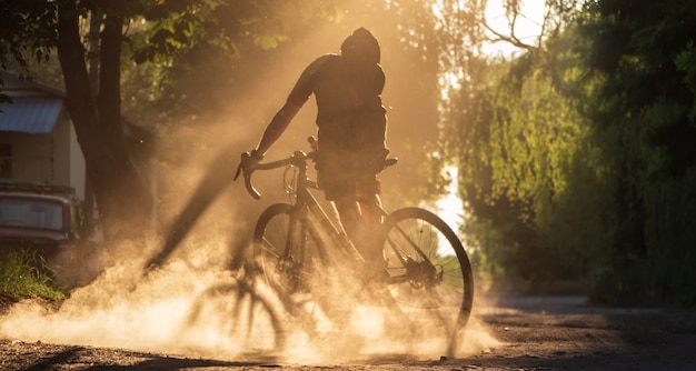 Cyclist riding a bicycle on a gravel road at sunset. A silhouette of young sporty man on a gravel bike in a cloud of dust.