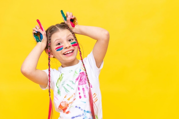 A dabbling girl stained in paints shows horns on a yellow isolated background The child is playing with colorful paints Courses for the development of children's creativity Happy childhood games