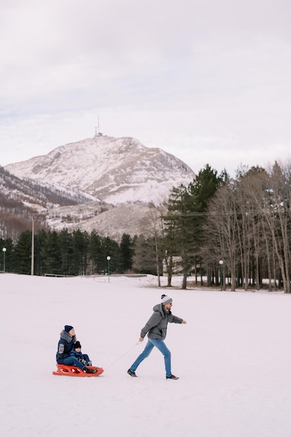 Dad carries mom and child on a sleigh across a snowy plain at the foot of the mountains