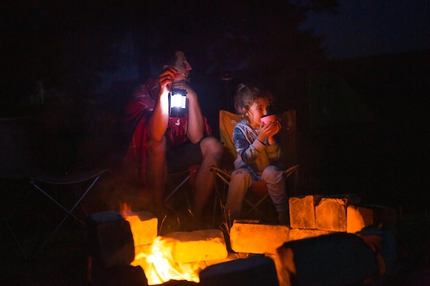 Dad and daughter sit at night by the fire in the open air in the summer in nature Family camping trip gatherings around the campfire Father's Day barbecue Camping lantern and tent