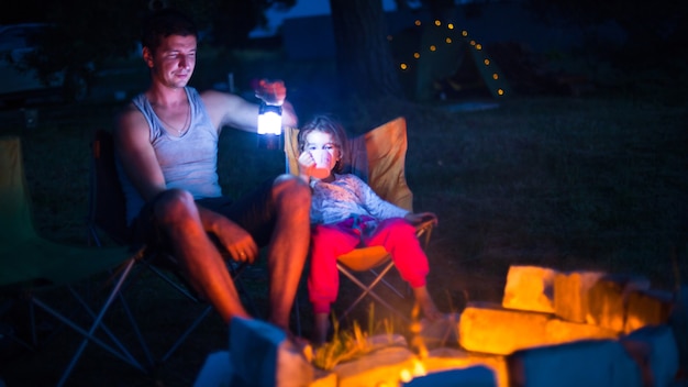 Dad and daughter sit at night by the fire in the open air in the summer in nature.