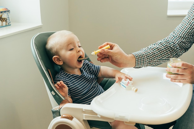 Dad father feeding cute baby with puree at home sitting at highchair Family food eating concept