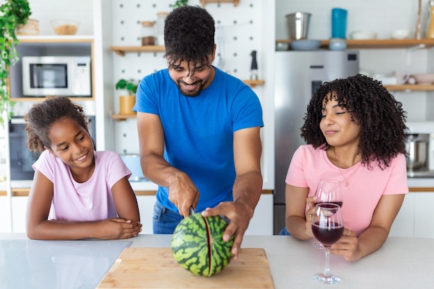 Dad is cutting watermelon while mom and daughter are watching him