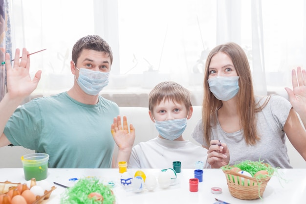 Dad, mom and child at the table in medical masks are painting Easter eggs for the holiday