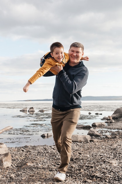 Dad plays with his son on the riverbank. A beautiful father and son are having fun playing and smiling on a sunny day. The child is in his father's arms. Outdoor recreation.