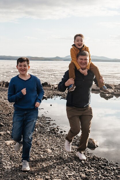 Dad plays with his sons on the riverbank. A beautiful father and two sons are having fun playing, running and smiling on a sunny day. The child is on his father's shoulders. Outdoor recreation.