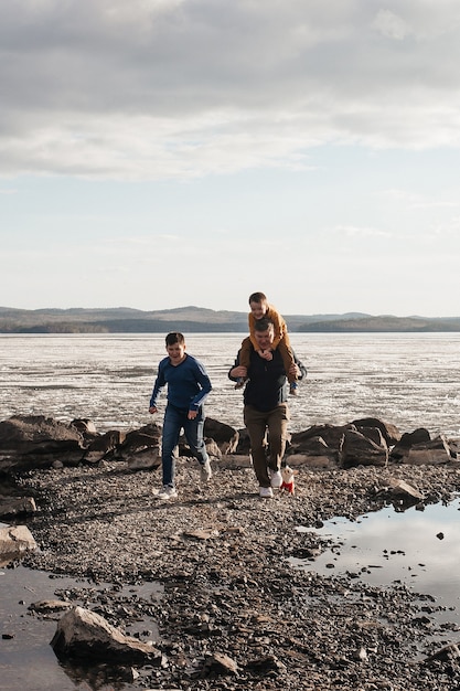 Dad plays with his sons on the riverbank. A beautiful father and two sons are having fun playing, running and smiling on a sunny day. The child is on his father's shoulders. Outdoor recreation.