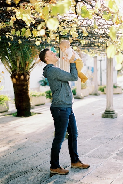 Dad raises a baby in his arms while standing in a pergola under the leaves