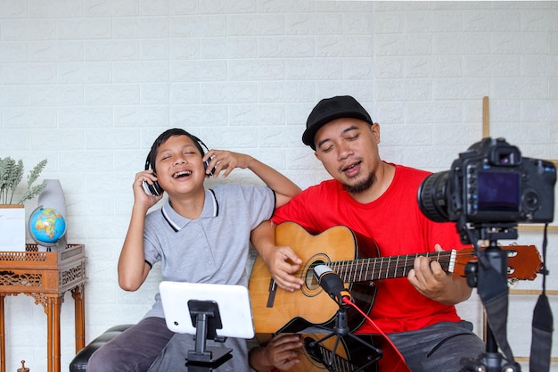 Dad and son singing and playing guitar together while doing video online streaming. Asian family spe