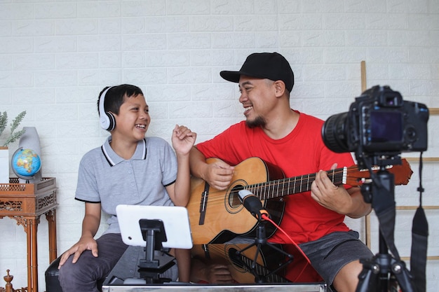 Dad and son singing and playing guitar together while doing video online streaming. Asian family spe