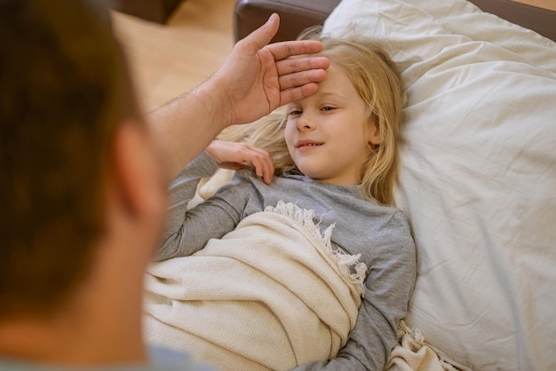 Dad touches the forehead of his sick child who is lying on the couch covered with a white blanket