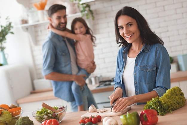 Daddy holding girl on the hands while wife cutting vegetables for salad and looking at the camera