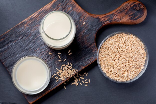 Dairy free oat milk in glass bottle and glass and oat seeds in glass bowl on wooden kitchen board on grey background