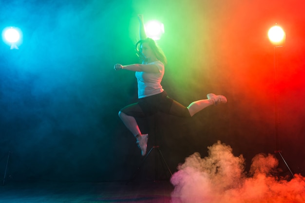 Dancing, sport, jazz funk and people concept - young woman jump in the darkness under colourful light.