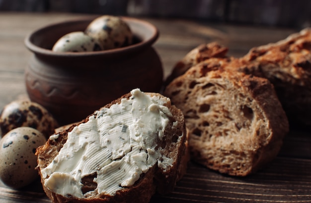 Dark buckwheat bread is spread with cottage cheese with herbs in a cut on a wooden table near quail eggs in a clay plate