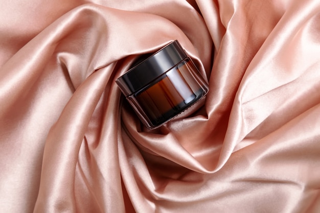 Dark glass jar of moisturizing cream in folds of silk pink cloth