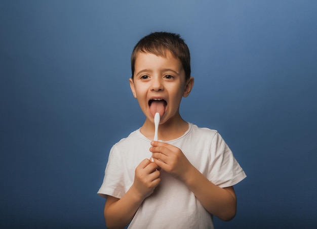 A dark-haired boy in a white T-shirt on a blue background brushes his teeth with a toothbrush. baby hygiene