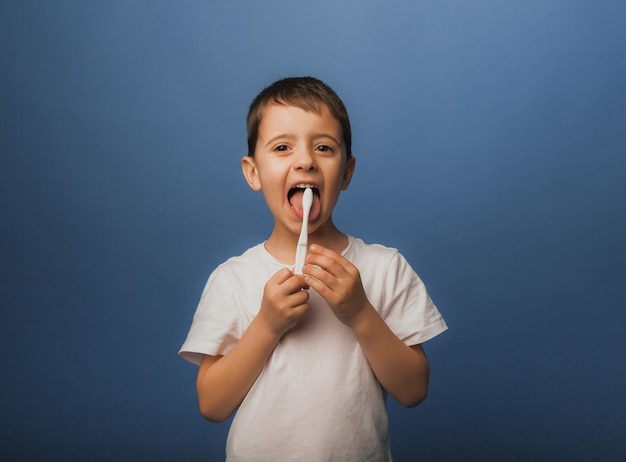 A dark-haired boy in a white T-shirt on a blue background brushes his teeth with a toothbrush. baby hygiene