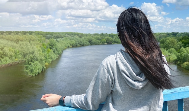 A dark haired caucasian girl stands with her back in a light gray jacket and looks towards the river and the forest in the city of Chernigiv in Ukraine.