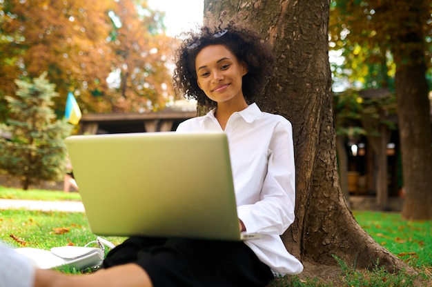 Dark-haired girl sitting under the tree with a laptop