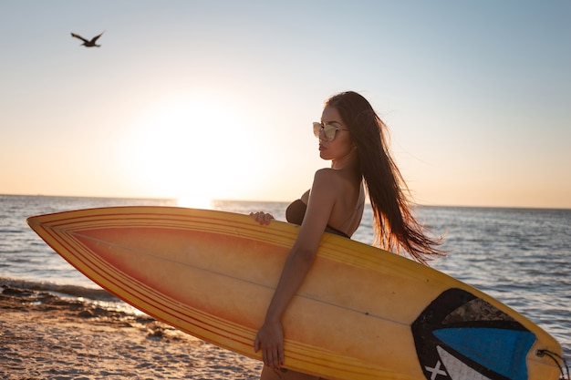 Dark-haired girl in a swimsuit and sunglasses walks on the sandy beach on the sunset and holds a surfboard .