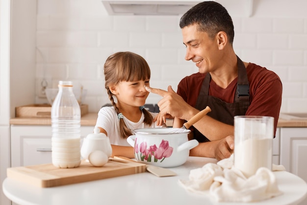 Dark haired man wearing maroon t shirt and apron cooking in kitchen with little daughter, father smeared the child's nose with flour, family spending happy time together.